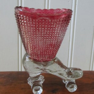 May include: A vintage, ruby red glass footed vase with a textured diamond pattern. The vase has a flared top and sits on a clear glass base with three rounded feet. The rim of the vase has a scalloped edge.