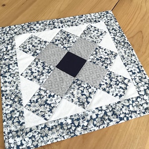 May include: Square quilted placemat with a floral pattern border. The center features a geometric design with white, gray, and navy blue fabric squares. The placemat is on a wooden surface.