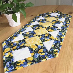 May include: A quilted table runner featuring a geometric pattern of alternating squares and triangles. The fabric displays a floral print with blue and yellow flowers on a dark background, interspersed with solid white fabric squares and triangles.