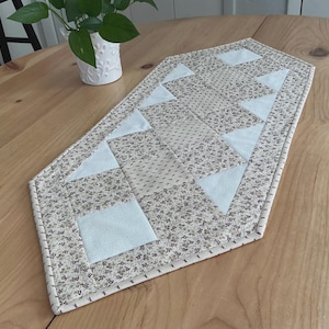 May include: A quilted table runner with a floral pattern in beige, gray, and white. The runner features light blue geometric shapes, including triangles and squares. The edges are finished with a contrasting stitch.