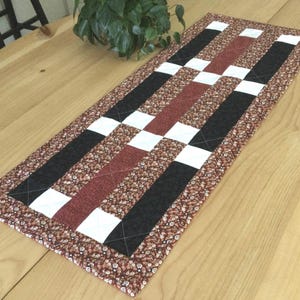 May include: A rectangular quilted table runner with a patchwork design. The runner features alternating stripes of black, white, and burgundy, with a floral patterned border. The runner is approximately 30 inches long.