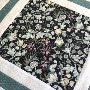 May include: A close-up of a quilted fabric with a floral pattern. The fabric is black with white, green, and pink flowers. The fabric is surrounded by a white border and a teal border.