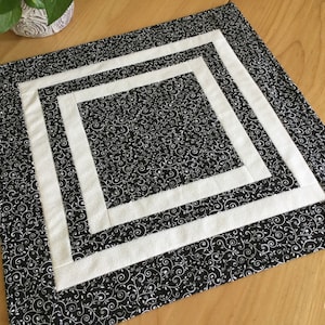 May include: A square quilted placemat with a black and white floral pattern. The placemat features two white square borders. The fabric has a repeating pattern of swirling vines and leaves. This decorative placemat is suitable for a dining table.