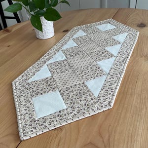 May include: A handmade table runner with a floral pattern in beige, gray, and light blue. The runner features geometric shapes, including triangles and squares, and is placed on a wooden table. A small white pot with a green plant is in the background.