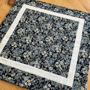 May include: A square quilted placemat with a floral pattern in navy, blue, and white. A white border frames the floral design. The placemat is placed on a wooden surface.