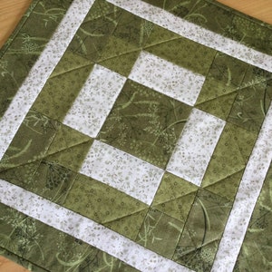 May include: A green and white quilted placemat with a floral pattern. The placemat has a white border and is made of fabric.