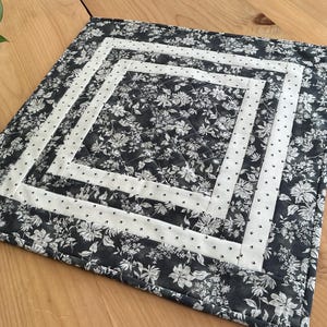 May include: A square quilted placemat with a floral pattern in shades of gray and white. The design features a central floral square, bordered by white bands with small black dots, and a floral outer edge. The placemat is on a wooden surface.