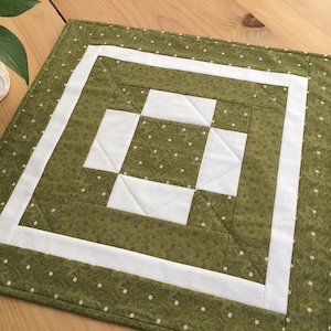 May include: A green and white quilted table runner with a geometric pattern. The runner has a white border and a green center with a white square in the middle.