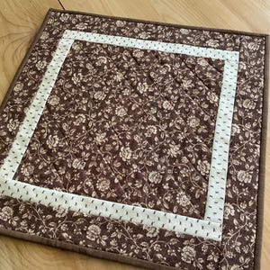 May include: A square, quilted placemat with a floral pattern in shades of brown and cream. A cream-colored border with small black dots frames the center. The edges are finished with a solid brown binding.
