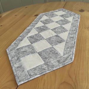 May include: A quilted table runner with a diamond pattern in shades of gray and white. The runner is placed on a wooden table. The edges are finished with a gray border.