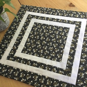 May include: A square table runner with a black background and a white floral pattern. The runner has a white border with a double-layered border.