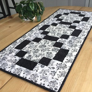 May include: Black and white checkered table runner with a floral pattern. The runner is made of fabric and has a gray border.