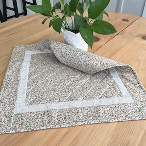 May include: A rectangular quilted placemat featuring a small-scale floral pattern in shades of green, red, and white. The fabric has a light-colored border and diagonal stitching lines throughout the surface, with one corner folded over to reveal the back.