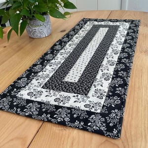 May include: A rectangular table runner with a floral pattern in black and white. The runner is made of fabric and has a quilted appearance. The design features alternating stripes of solid black, white with small black flowers, and black with white floral patterns.