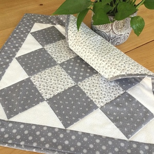 May include: A gray and white checkered table runner with a white border and a gray floral pattern. The runner is folded in half and is lying on a wooden table.