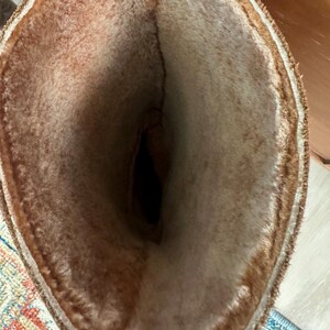 Puede incluir: Una bota alta con una abertura ancha, que presenta un forro de piel sint&eacute;tica grueso y afelpado de color marr&oacute;n claro que se extiende hasta el borde superior. El exterior de la bota es de un material marr&oacute;n oscuro y el interior muestra una cavidad.