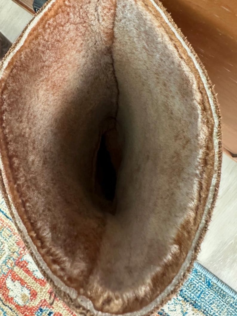 Puede incluir: Una bota alta con una abertura ancha, que presenta un forro de piel sint&eacute;tica grueso y afelpado de color marr&oacute;n claro que se extiende hasta el borde superior. El exterior de la bota es de un material marr&oacute;n oscuro y el interior muestra una cavidad.
