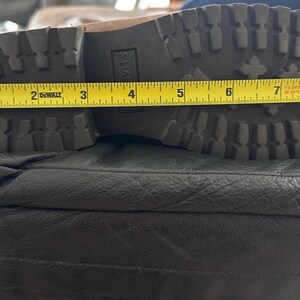 Puede incluir: Una suela de bota marr&oacute;n con un patr&oacute;n de tacos profundos y un tac&oacute;n de bloque. Una cinta m&eacute;trica amarilla DeWalt est&aacute; colocada sobre la suela, indicando una longitud de aproximadamente 23 cm desde el tal&oacute;n hasta la punta de la bota.