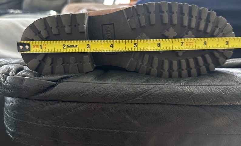 Puede incluir: Una suela de bota marr&oacute;n con un patr&oacute;n de tacos profundos y un tac&oacute;n de bloque. Una cinta m&eacute;trica amarilla DeWalt est&aacute; colocada sobre la suela, indicando una longitud de aproximadamente 23 cm desde el tal&oacute;n hasta la punta de la bota.