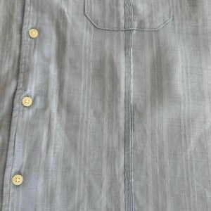 Puede incluir: Camisa azul claro con botones y bolsillo en el pecho. La camisa tiene detalles de rayas verticales y botones color crema. La tela parece ser un material ligero y texturizado.