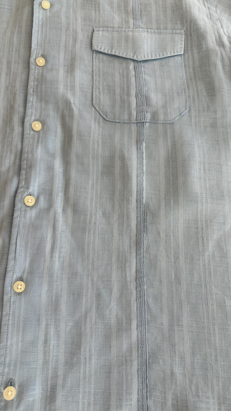 Puede incluir: Camisa azul claro con botones y bolsillo en el pecho. La camisa tiene detalles de rayas verticales y botones color crema. La tela parece ser un material ligero y texturizado.