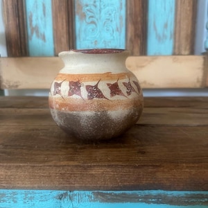 May include: A small, round ceramic pot with a wide opening and a short neck. The exterior features a cream-colored upper section, a central band with repeating dark brown leaf-like motifs, and a textured, dark brown base with a weathered finish.
