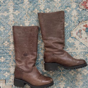 Puede incluir: Un par de botas de cuero marr&oacute;n a media pantorrilla con acabado desgastado, que presentan una suela gruesa de tacos negra y un tac&oacute;n bajo. El nombre de la marca Shabbies Amsterdam est&aacute; grabado en el lateral de la ca&ntilde;a junto a un logo de corona.