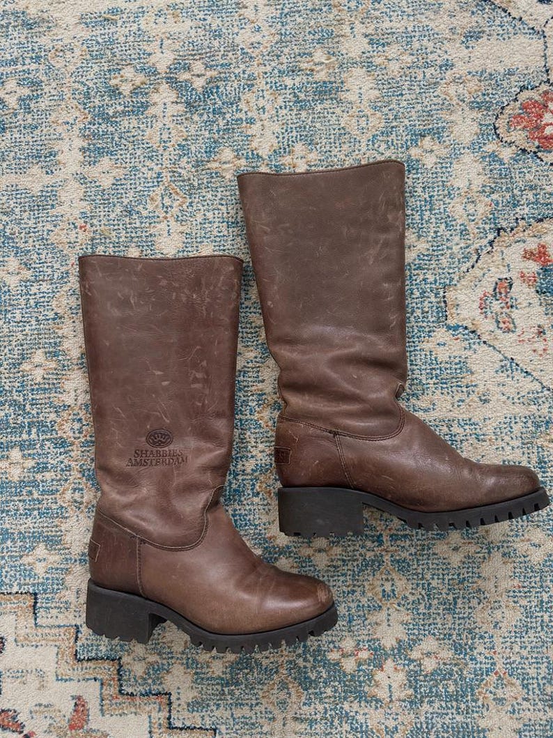 Puede incluir: Un par de botas de cuero marr&oacute;n a media pantorrilla con acabado desgastado, que presentan una suela gruesa de tacos negra y un tac&oacute;n bajo. El nombre de la marca Shabbies Amsterdam est&aacute; grabado en el lateral de la ca&ntilde;a junto a un logo de corona.