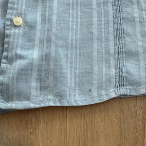 Puede incluir: Primer plano de una camisa abotonada azul claro con un patr&oacute;n de cuadros. La camisa tiene botones blancos y costuras verticales en el lado derecho. La tela parece ser ligera, adecuada para climas c&aacute;lidos.