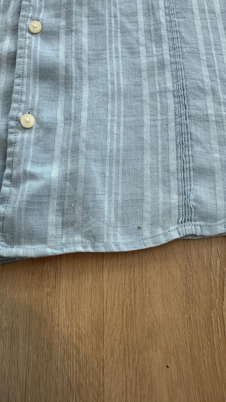 Puede incluir: Primer plano de una camisa abotonada azul claro con un patr&oacute;n de cuadros. La camisa tiene botones blancos y costuras verticales en el lado derecho. La tela parece ser ligera, adecuada para climas c&aacute;lidos.