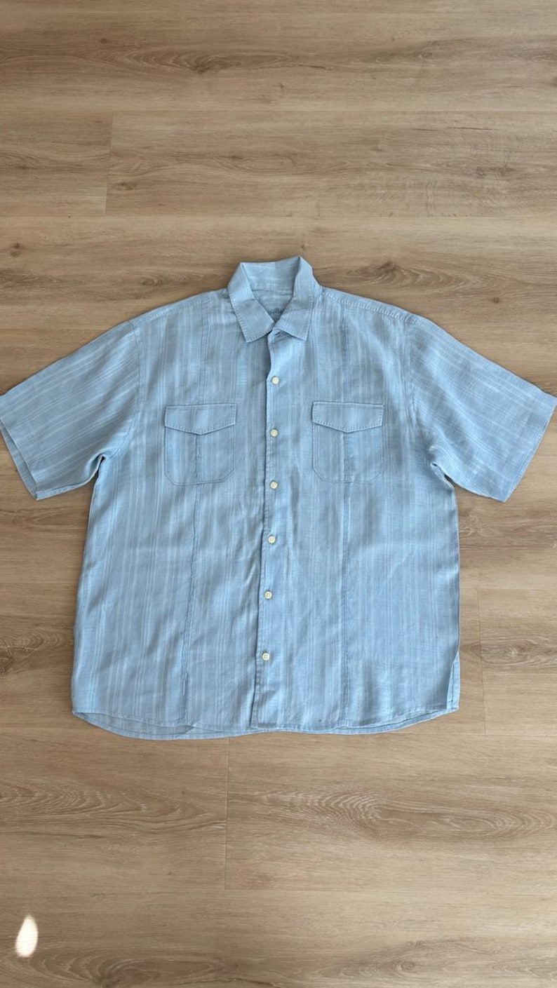 Puede incluir: Camisa azul claro de manga corta con estampado de rayas verticales. La camisa tiene dos bolsillos en el pecho y una botonadura frontal. El cuello es un cuello cl&aacute;sico. La camisa est&aacute; hecha de tela ligera.