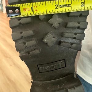 Puede incluir: Una suela de bota de color marr&oacute;n oscuro con un patr&oacute;n de tacos profundos y el nombre de la marca SHABBIES grabado en el centro. Una cinta m&eacute;trica amarilla DeWalt se coloca a trav&eacute;s del ancho de la suela, mostrando una medida de 8,25 cm.