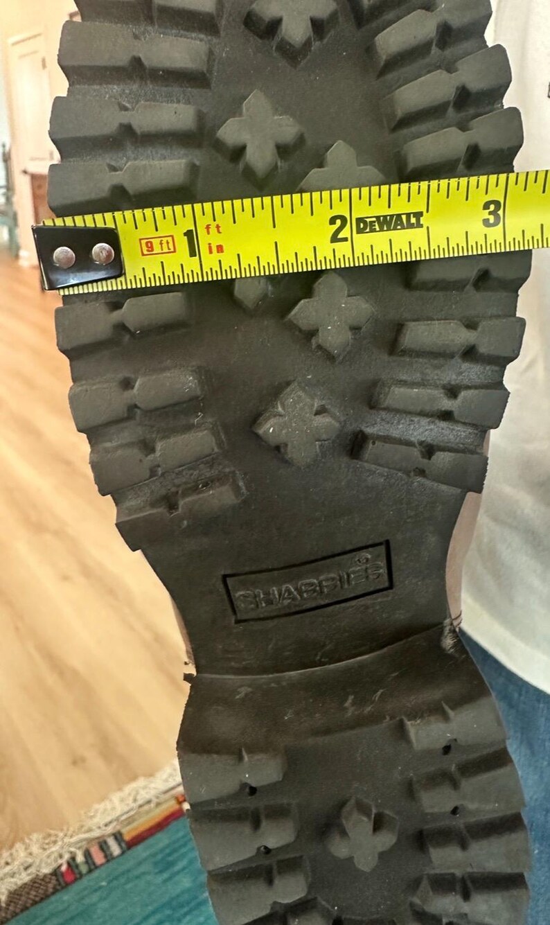 Puede incluir: Una suela de bota de color marr&oacute;n oscuro con un patr&oacute;n de tacos profundos y el nombre de la marca SHABBIES grabado en el centro. Una cinta m&eacute;trica amarilla DeWalt se coloca a trav&eacute;s del ancho de la suela, mostrando una medida de 8,25 cm.