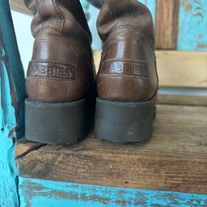 Puede incluir: Un par de botas de cuero marr&oacute;n con altura a media pantorrilla, una suela gruesa de goma marr&oacute;n oscuro y el nombre de la marca SHABBIES grabado en un parche de cuero rectangular en la parte posterior de cada tal&oacute;n.