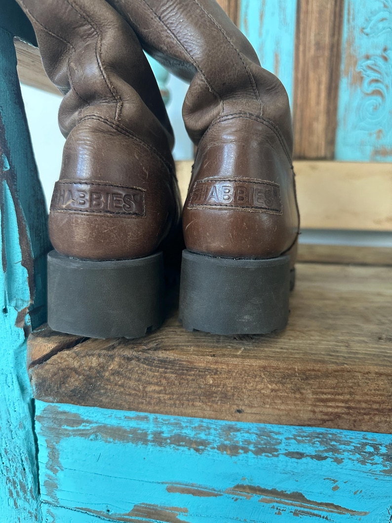 Puede incluir: Un par de botas de cuero marr&oacute;n con altura a media pantorrilla, una suela gruesa de goma marr&oacute;n oscuro y el nombre de la marca SHABBIES grabado en un parche de cuero rectangular en la parte posterior de cada tal&oacute;n.