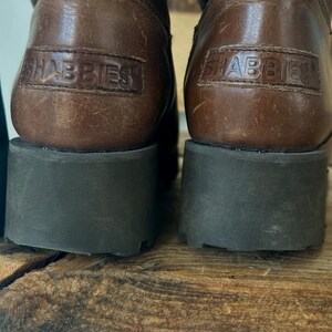 Puede incluir: Un par de botas de cuero marr&oacute;n con una suela gruesa de goma gris oscuro. La parte posterior de cada tal&oacute;n muestra un parche de cuero rectangular grabado con el nombre de la marca SHABBIES en letras may&uacute;sculas. El cuero tiene una textura natural.