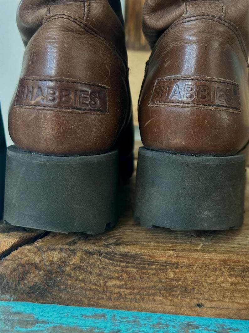 Puede incluir: Un par de botas de cuero marr&oacute;n con una suela gruesa de goma gris oscuro. La parte posterior de cada tal&oacute;n muestra un parche de cuero rectangular grabado con el nombre de la marca SHABBIES en letras may&uacute;sculas. El cuero tiene una textura natural.