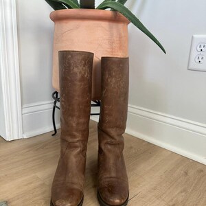 Puede incluir: Un par de botas altas de cuero marr&oacute;n con acabado desgastado, que presentan punteras redondeadas, ca&ntilde;a lisa y suelas gruesas de tacos en color marr&oacute;n oscuro. Las botas muestran rozaduras visibles y variaciones naturales en la textura del cuero.