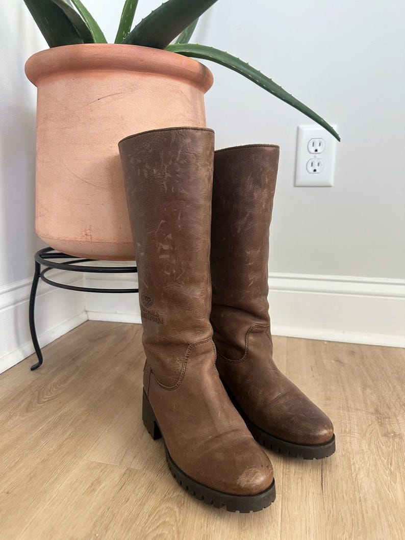 Puede incluir: Un par de botas de cuero marr&oacute;n a media pantorrilla con acabado desgastado, punta redondeada y suela gruesa de tacos en color marr&oacute;n oscuro. Las botas presentan un logotipo sutil grabado en el lado exterior de la ca&ntilde;a y un tac&oacute;n bajo y cuadrado.