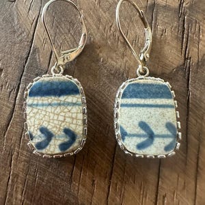 May include: A pair of dangle earrings featuring rectangular ceramic shards with a cream-colored crackle glaze, decorated with blue horizontal stripes and leaf-like patterns, set in silver-toned metal frames with lever-back closures.