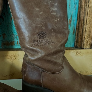 Puede incluir: Una bota alta de cuero marr&oacute;n con un acabado desgastado y una suela gruesa de tacos negros. La ca&ntilde;a de la bota presenta el logotipo y el texto SHABBIES AMSTERDAM en relieve. El cuero muestra pliegues naturales y desgaste en la superficie.