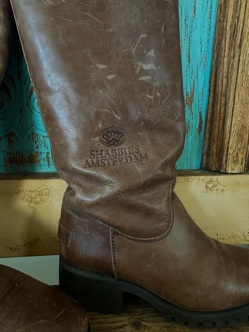 Puede incluir: Una bota alta de cuero marr&oacute;n con un acabado desgastado y una suela gruesa de tacos negros. La ca&ntilde;a de la bota presenta el logotipo y el texto SHABBIES AMSTERDAM en relieve. El cuero muestra pliegues naturales y desgaste en la superficie.