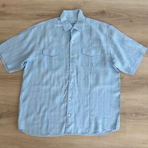 Puede incluir: Camisa azul claro de manga corta con dos bolsillos en el pecho y cierre de botones. La camisa tiene un patr&oacute;n de rayas verticales y un cuello cl&aacute;sico. La prenda est&aacute; hecha de una tela ligera, adecuada para climas c&aacute;lidos.