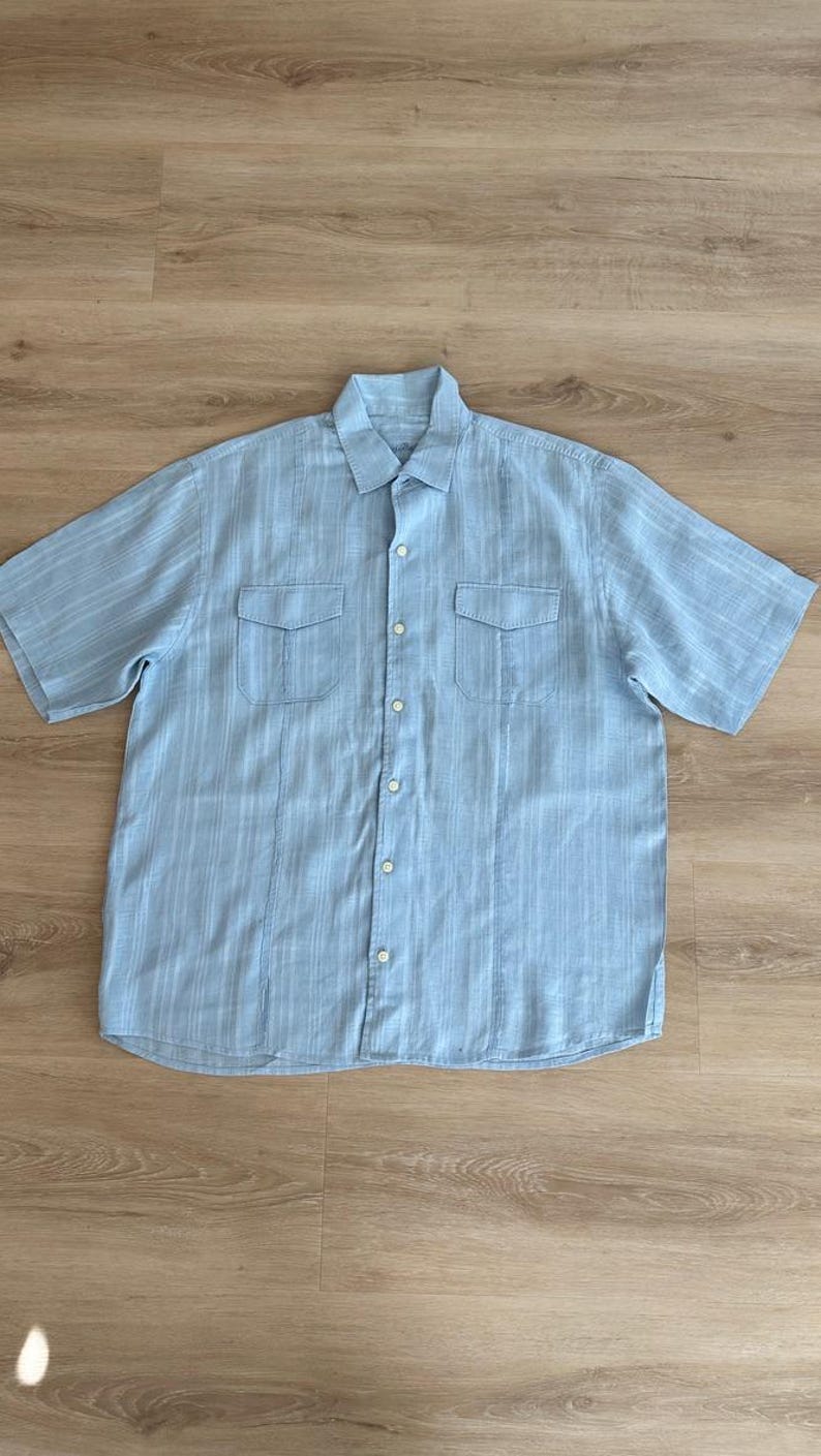 Puede incluir: Camisa azul claro de manga corta con dos bolsillos en el pecho y cierre de botones. La camisa tiene un patr&oacute;n de rayas verticales y un cuello cl&aacute;sico. La prenda est&aacute; hecha de una tela ligera, adecuada para climas c&aacute;lidos.