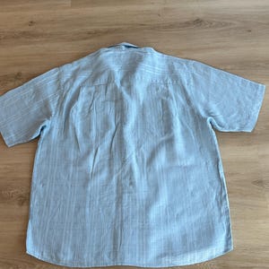 Puede incluir: Camisa azul claro de manga corta con un patr&oacute;n de rayas verticales. La camisa tiene cuello abotonado y un corte holgado. La parte trasera de la camisa es visible, mostrando el canes&uacute; y las costuras.