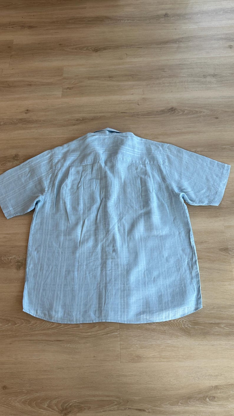 Puede incluir: Camisa azul claro de manga corta con un patr&oacute;n de rayas verticales. La camisa tiene cuello abotonado y un corte holgado. La parte trasera de la camisa es visible, mostrando el canes&uacute; y las costuras.