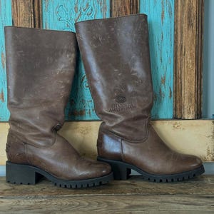 Puede incluir: Un par de botas de cuero marr&oacute;n a media pantorrilla con tac&oacute;n de bloque y suela de goma gruesa con tacos. Las botas tienen un acabado desgastado y el texto SHABBIES AMSTERDAM grabado en el lado exterior de la ca&ntilde;a cerca del &aacute;rea del tobillo.