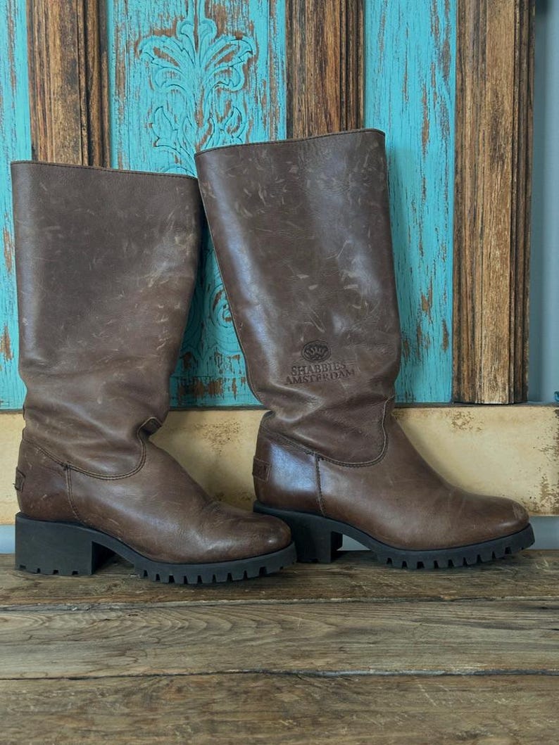 Puede incluir: Un par de botas de cuero marr&oacute;n a media pantorrilla con tac&oacute;n de bloque y suela de goma gruesa con tacos. Las botas tienen un acabado desgastado y el texto SHABBIES AMSTERDAM grabado en el lado exterior de la ca&ntilde;a cerca del &aacute;rea del tobillo.