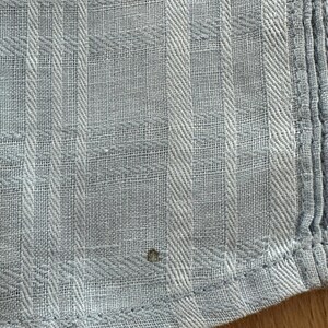 Puede incluir: Primer plano de una tela azul claro con un patr&oacute;n de rayas texturizado. La tela tiene una apariencia tejida con l&iacute;neas verticales y horizontales que crean un dise&ntilde;o similar a una cuadr&iacute;cula. El borde de la tela es visible, mostrando un dobladillo cosido.