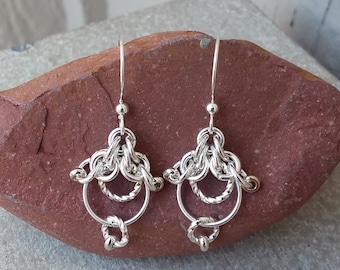 Peut inclure: Une paire de boucles d'oreilles en cotte de mailles argentées avec un design unique, présentant des motifs tissés complexes et des éléments circulaires. Les boucles d'oreilles sont présentées sur une surface en pierre brun-rouge. Le texte "Maillewerks Chainmaille Jewelry by Hanan Hall" est visible.